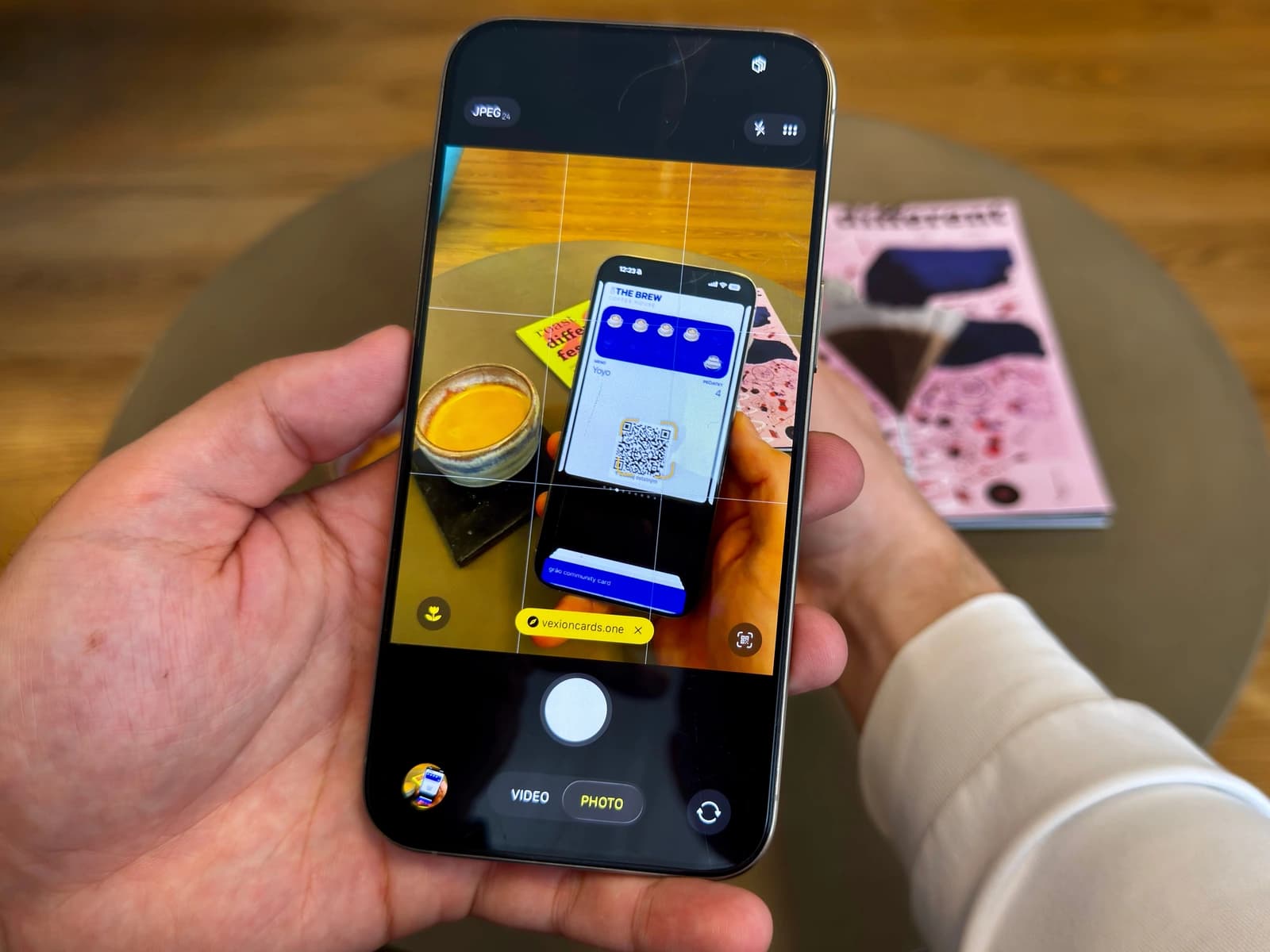 A hand holding a smartphone with the camera app open, scanning a QR code on a digital café loyalty card shown on another phone on a table next to an espresso and magazines.