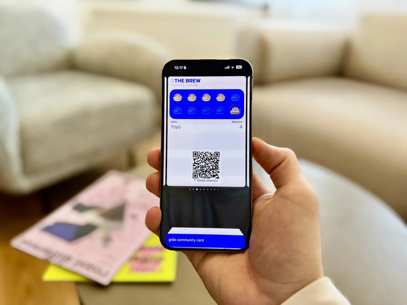 A hand holding a smartphone showing a digital café loyalty card with stamp icons and a QR code, with a blurred table of magazines in the background.